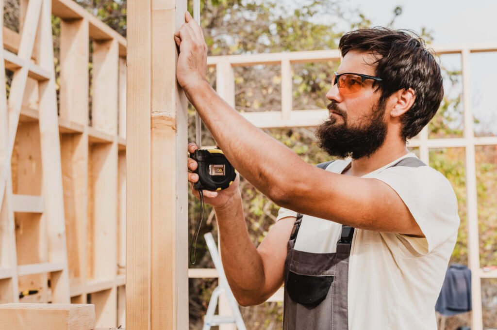 Exterior Services Exterior home services contractor measuring wooden framing during outdoor construction project, wearing safety glasses and overalls.